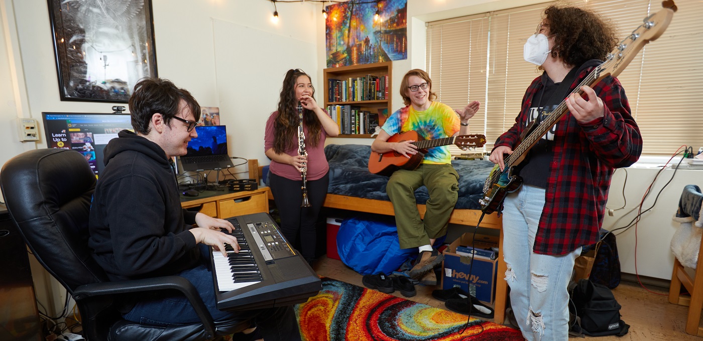 4 students with instruments (keyboard, guitar, clarinet, bass) hang out in a desk chair and on a bed in a PLU residence hall room.