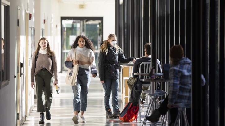 Students wearing backpacks walk down the brightly lit hallway of the PLU Morken Center for Learning & Technology. Next to the floor length windows, other students sit at tables studying.
