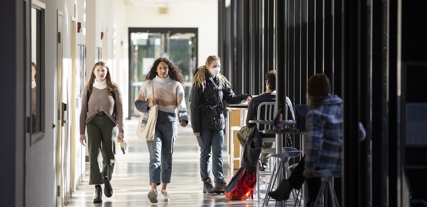 Students wearing backpacks walk down the brightly lit hallway of the PLU Morken Center for Learning & Technology. Next to the floor length windows, other students sit at tables studying.
