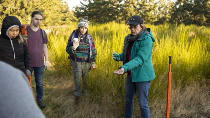 Students and a professor stand a field. The professor is holding their hand out showing students something. Students and professor are wearing hats and jackets.