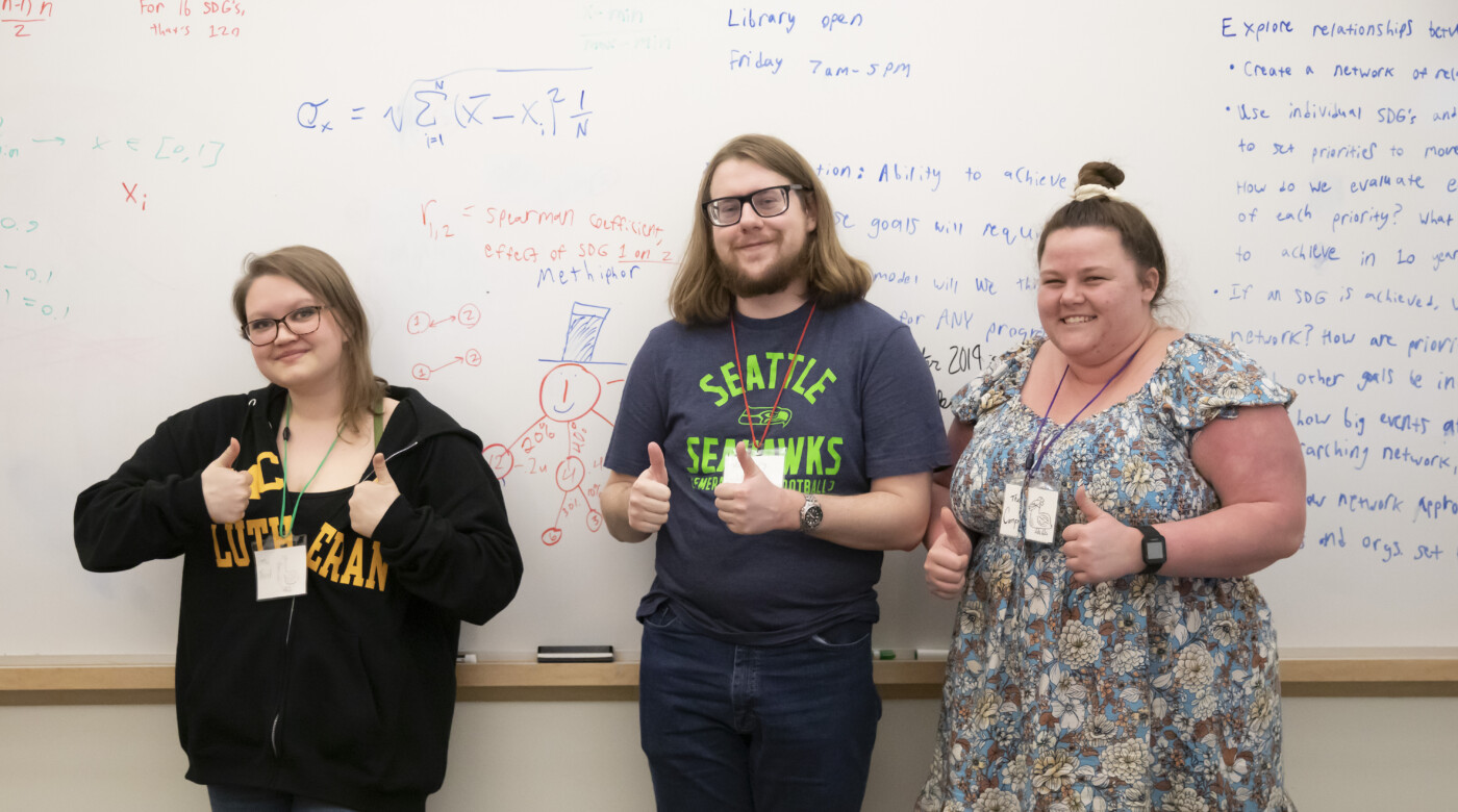 Three PLU students give the camera a thumbs up. They are standing in front of a white board that has math formulas on it.