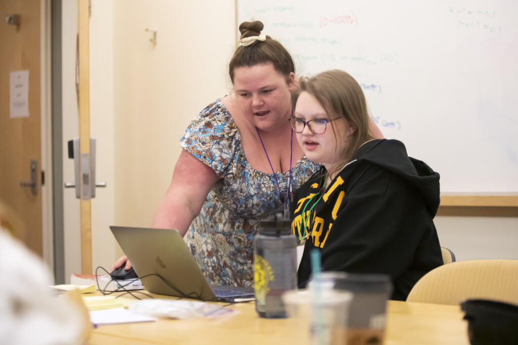 Two PLU students look at a lap top. One student is sitting and the other is looking over their shoulder at the computer.