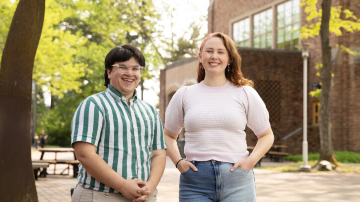 Two PLU students smile into the camera. The student on the left is wearing a green and white stripped shirt and glasses and has short dark hair. The student on the right it wearing a pink short sleeved shirt and has shoulder length red hair.