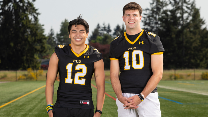 Two PLU football players wearing their jerseys smile into the camera while standing on the practice football field.