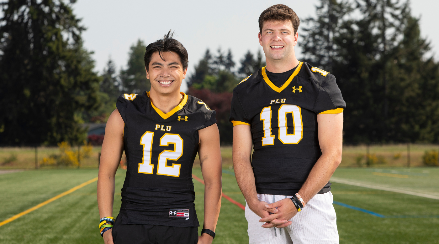 Two PLU football players wearing their jerseys smile into the camera while standing on the practice football field.