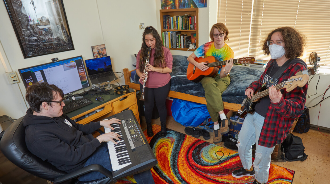 four students play different musical instruments: a keyboard, clarinet, guitar, and bass. They are standing and sitting on a colorful rainbow rug in a dorm room.