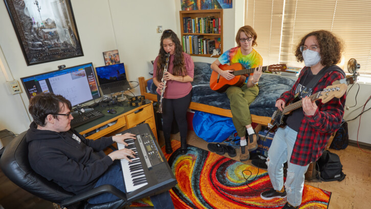 four students play different musical instruments: a keyboard, clarinet, guitar, and bass. They are standing and sitting on a colorful rainbow rug in a dorm room.