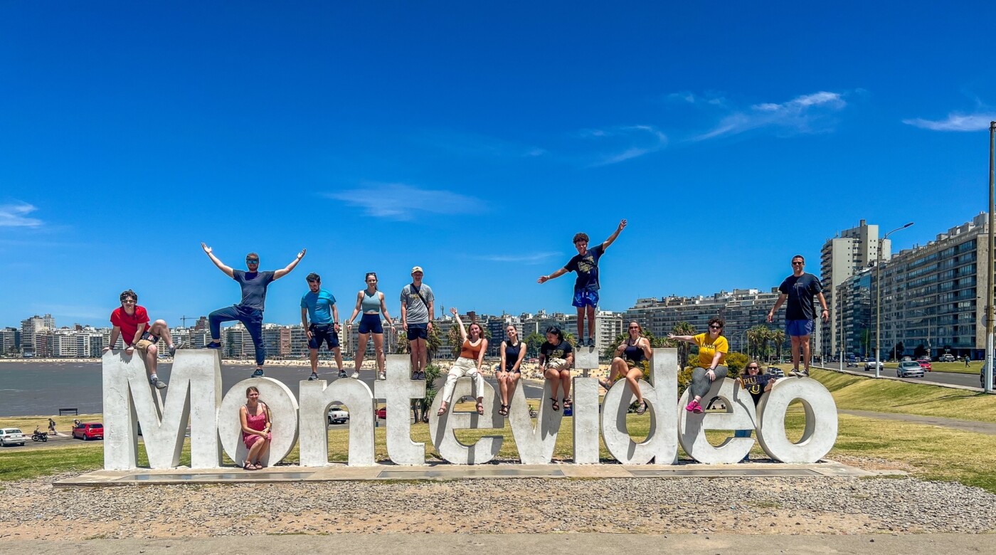 Students sit on a large sign that says Montevideo. The sun it out and the sky is blue.