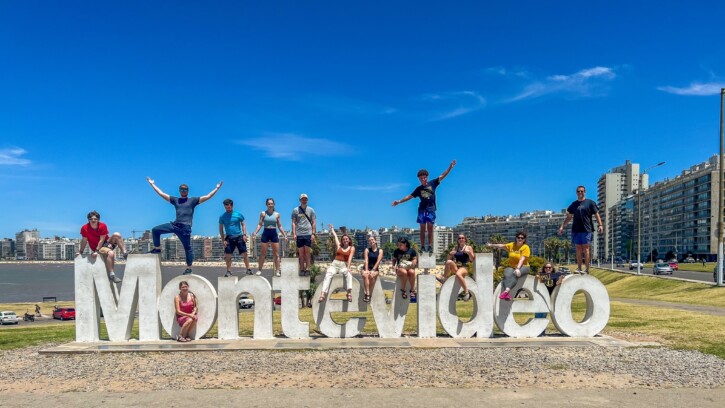 Students sit on a large sign that says Montevideo. The sun it out and the sky is blue.