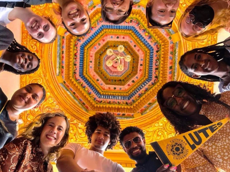 Students look down into the camera they are standing in a circle and there is a ornate golden ceiling above them.