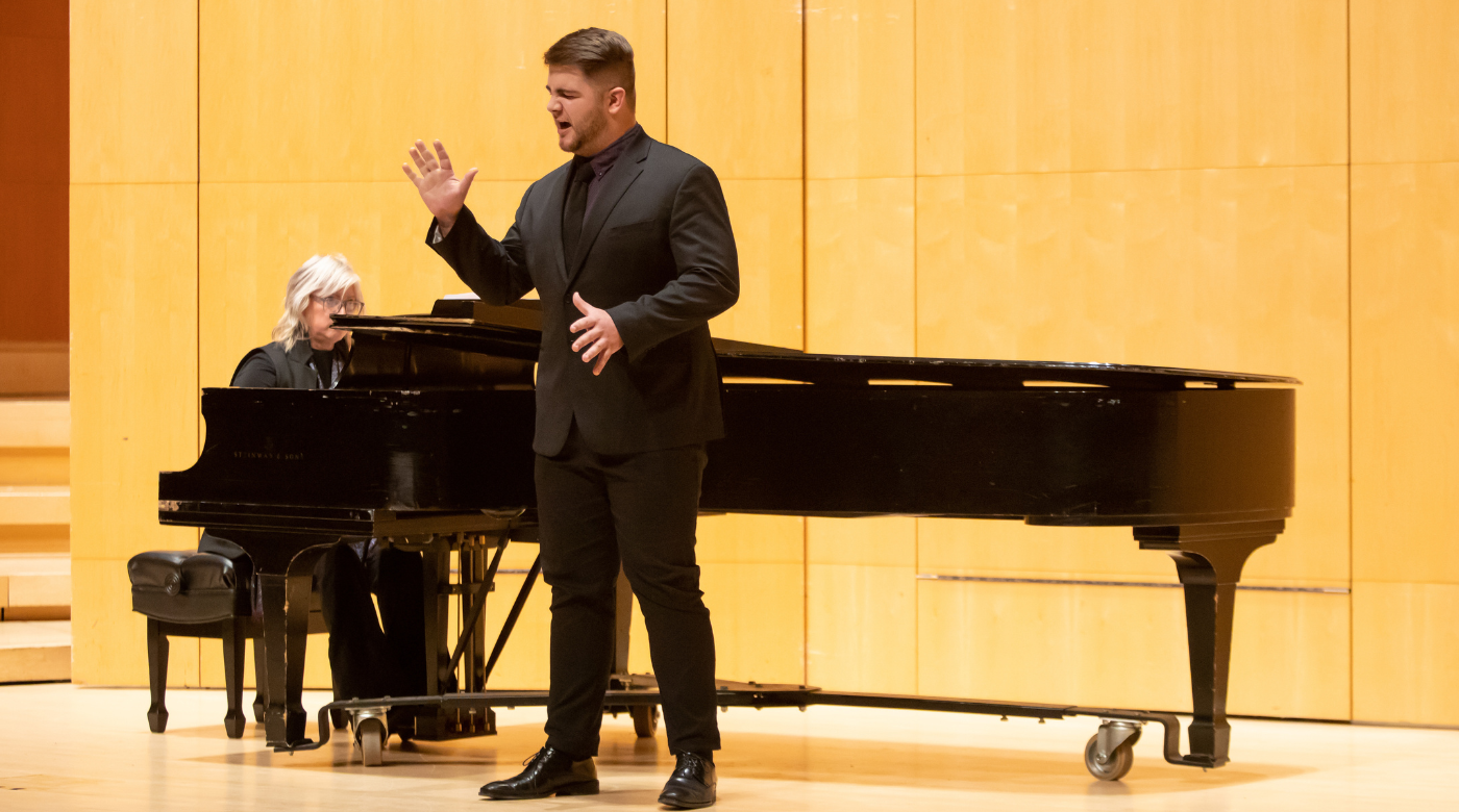 PLU student sings in front of a piano played by another person. The singer is wearing an all black suit.