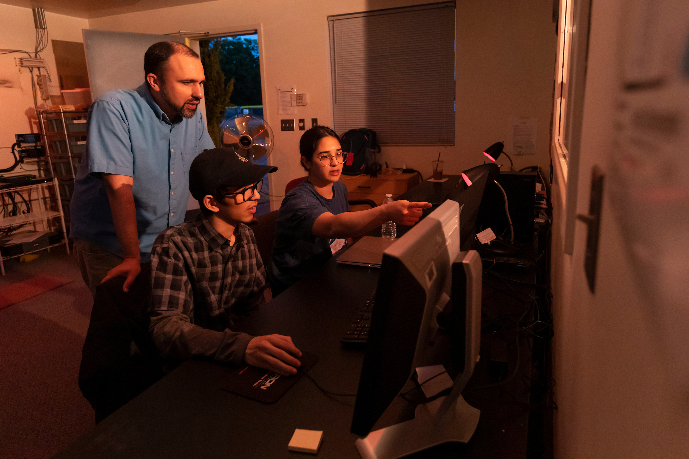 Three people stand next to a computer. One of the students points to the computer screen.