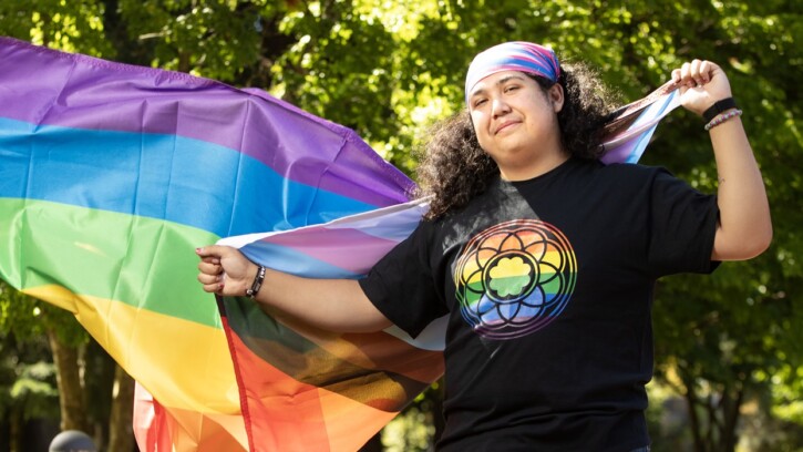 A student wearing a bandana and a black shirt with the PLU rose window in rainbow flag colors holds a pride flag that is flowing behind them.