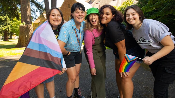 A group of 5 students stand together outside on the PLU campus, slightly crouched and smiling at the camera. One is holding a small pride flag and another has a large pride flag wrapped around their shoulders.