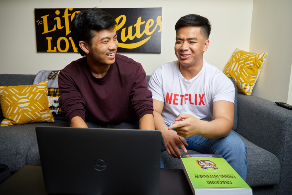 Two students look at a computer while sitting on a couch. One is wearing a Netflix shirt.
