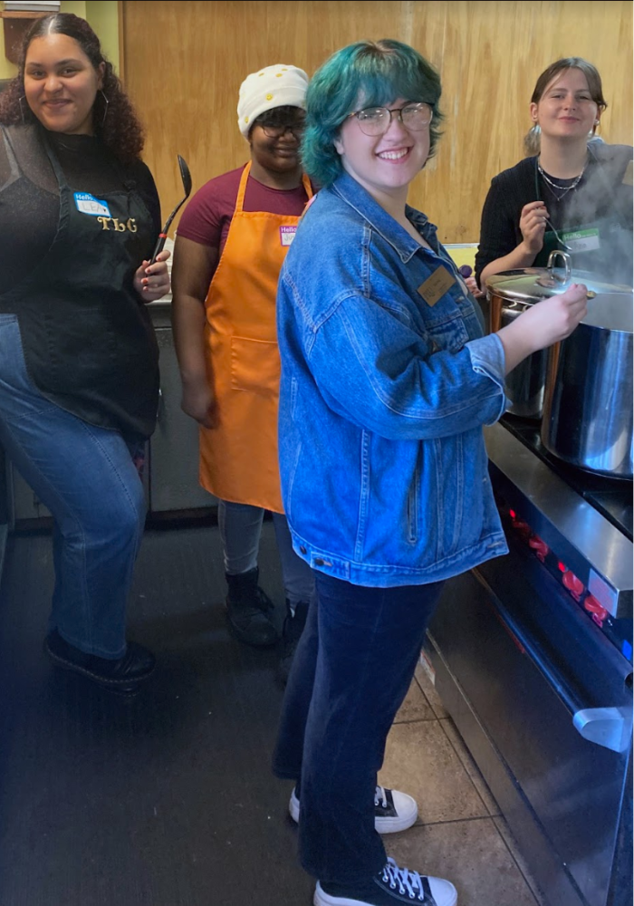 Four people look into the camera while cooking a pot of soup on the stove.