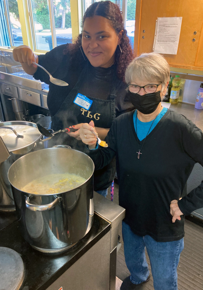 Two people look into the camera and hold up spoons of soup. There is a big pan of soup on the stove next to them.