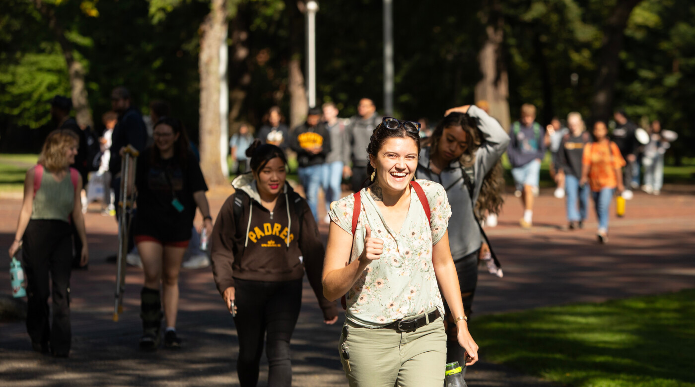 Students walk across campus. A student in the center of the photo smiles into the camera and gives a thumbs up.