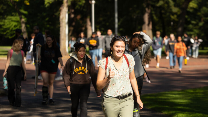 Students walk across campus. A student in the center of the photo smiles into the camera and gives a thumbs up.