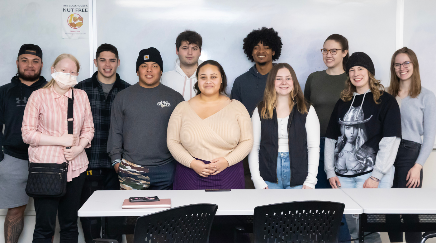PLU communications class stands together in front of white board and looks into the camera.