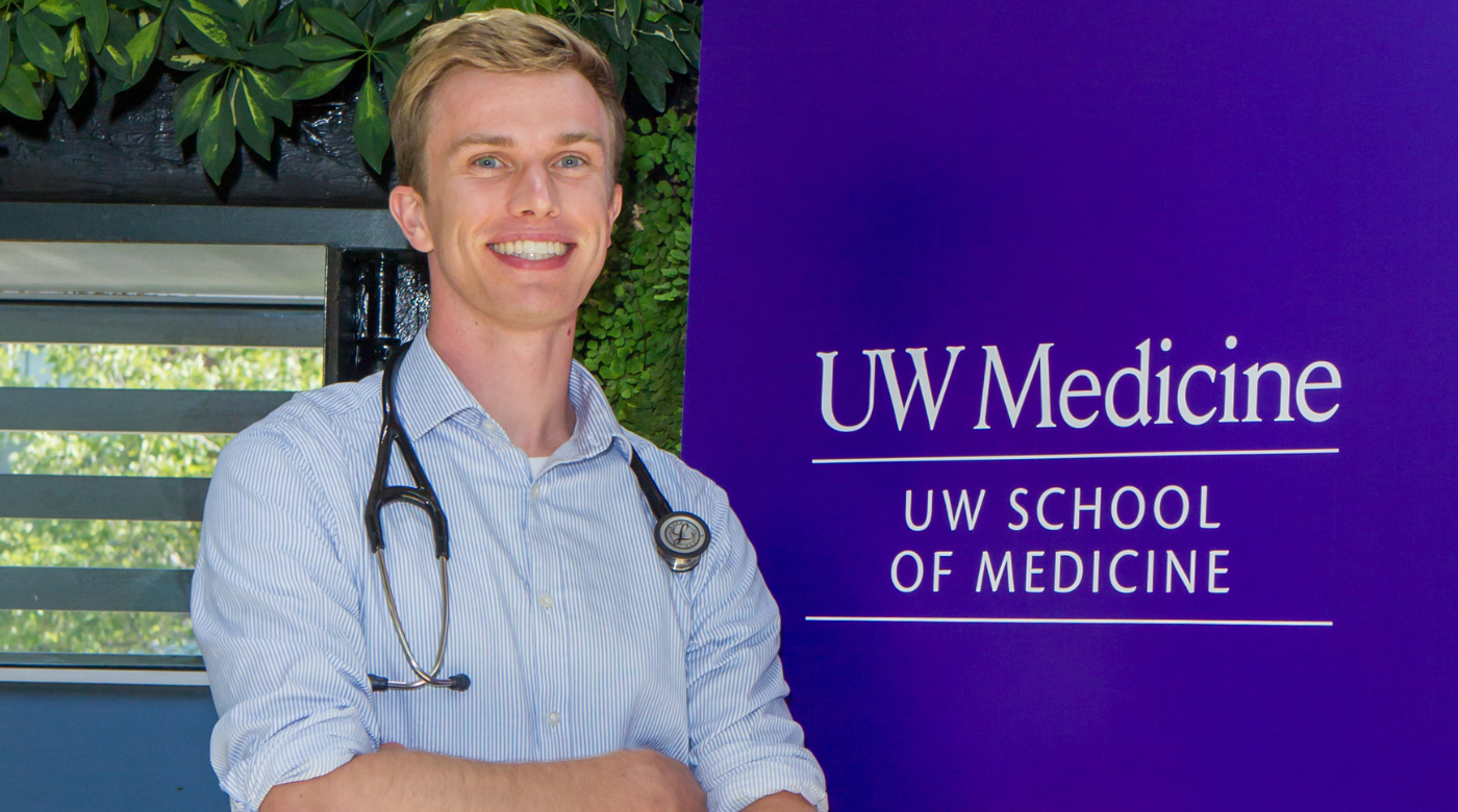 Student folds their arms and smiles into the camera and is standing in front of a purple UW Medicine sign.