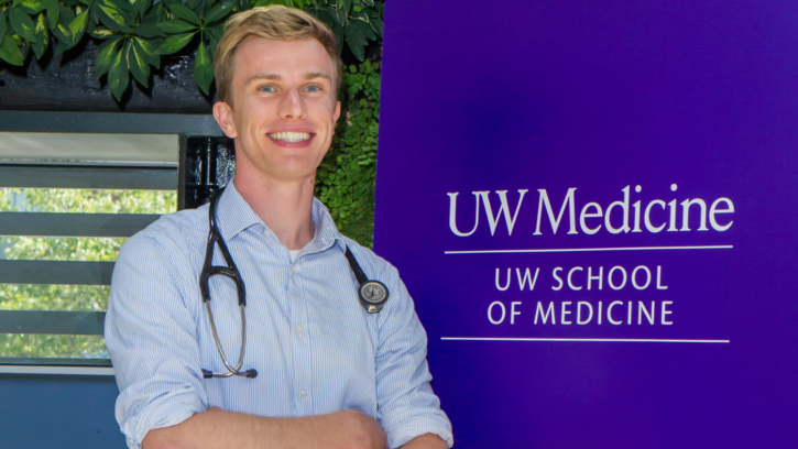 Student folds their arms and smiles into the camera and is standing in front of a purple UW Medicine sign.