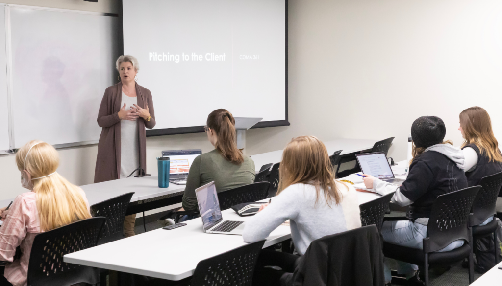 A professor stands in front of a classroom of students. There is a powerpoint presentation behind her that reads "How to pitch a client"