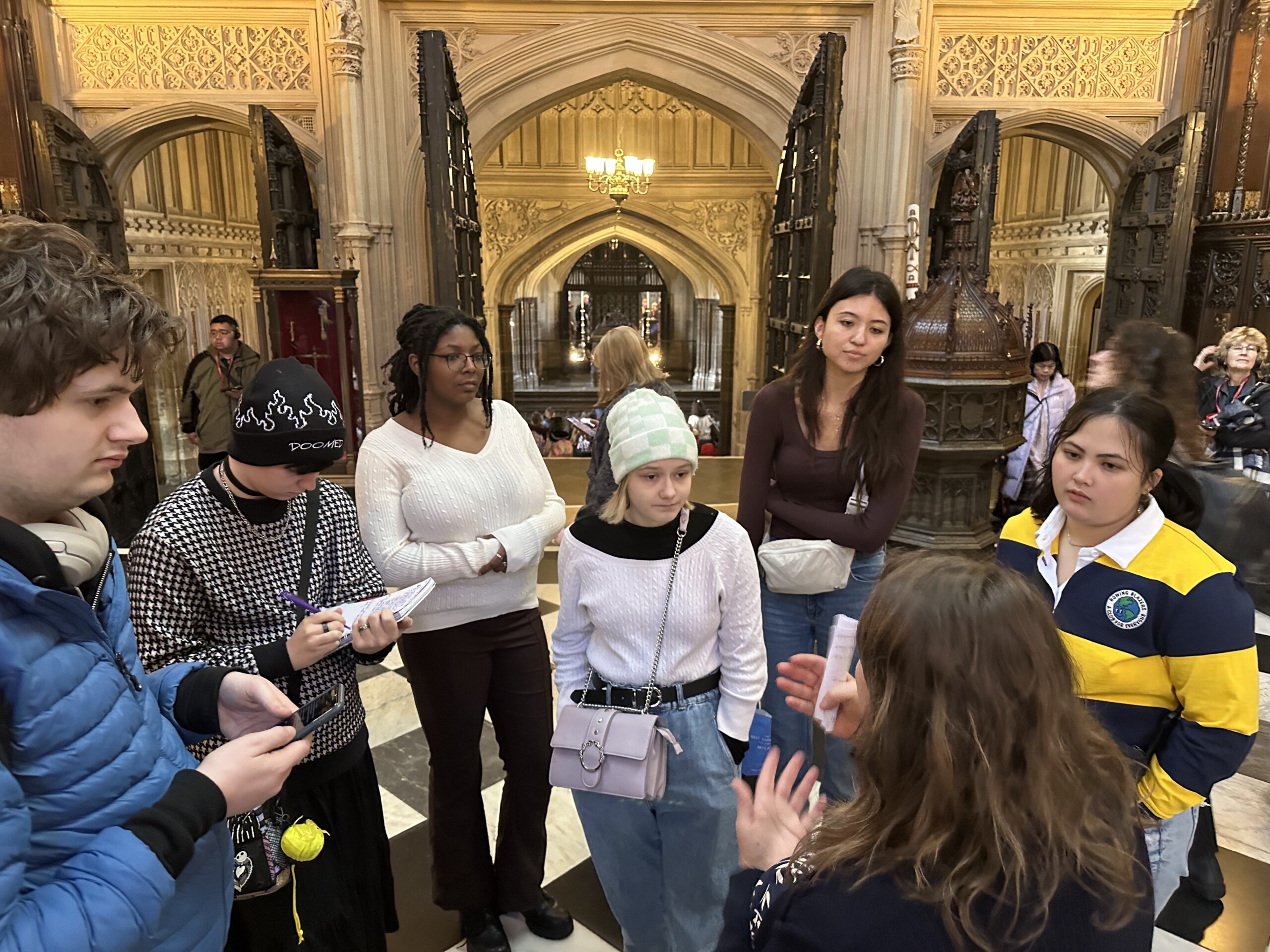 The back of a tour guide faces the camera. Students face the tour guide, listening to information about Westminster Abbey.