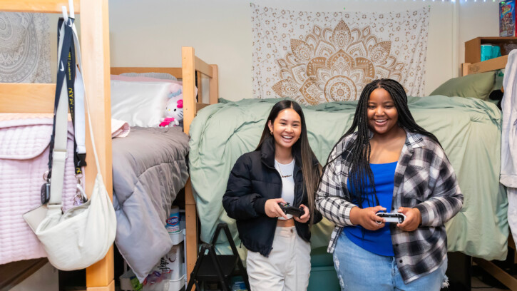 Two students look into the camera while they stand playing video games.