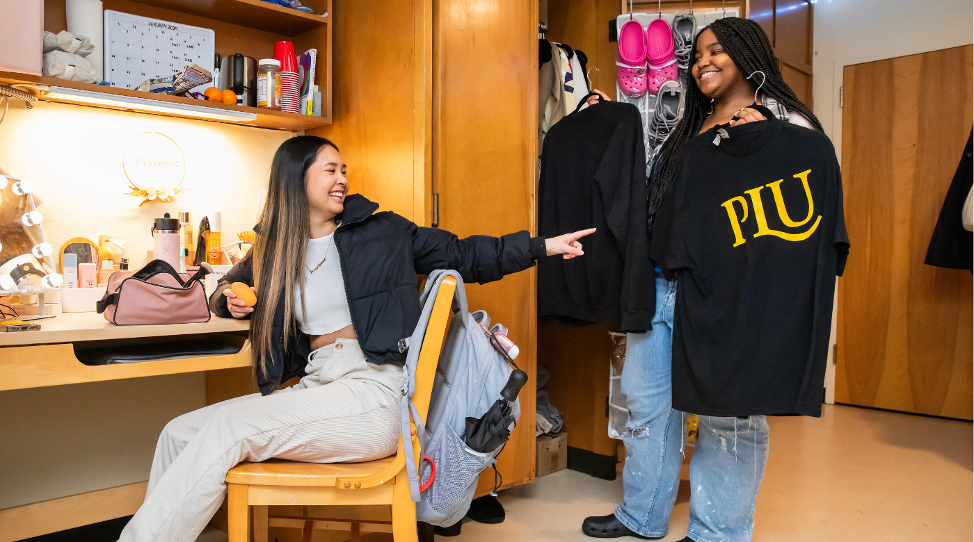 One students sits in their dorm room and another holds a PLU shirt in front of the closet.