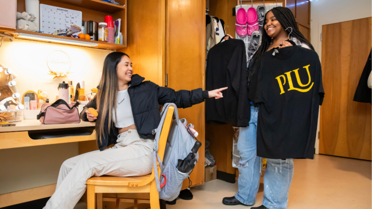 One students sits in their dorm room and another holds a PLU shirt in front of the closet.