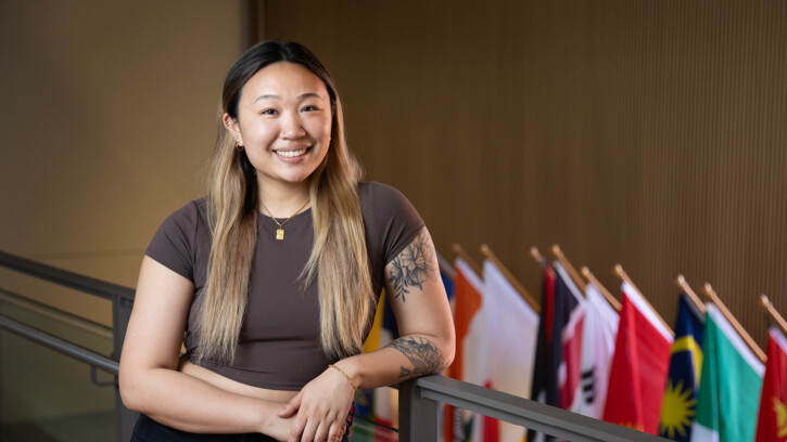 Student leans on a banister and smiles into the camera, flags are in the background.