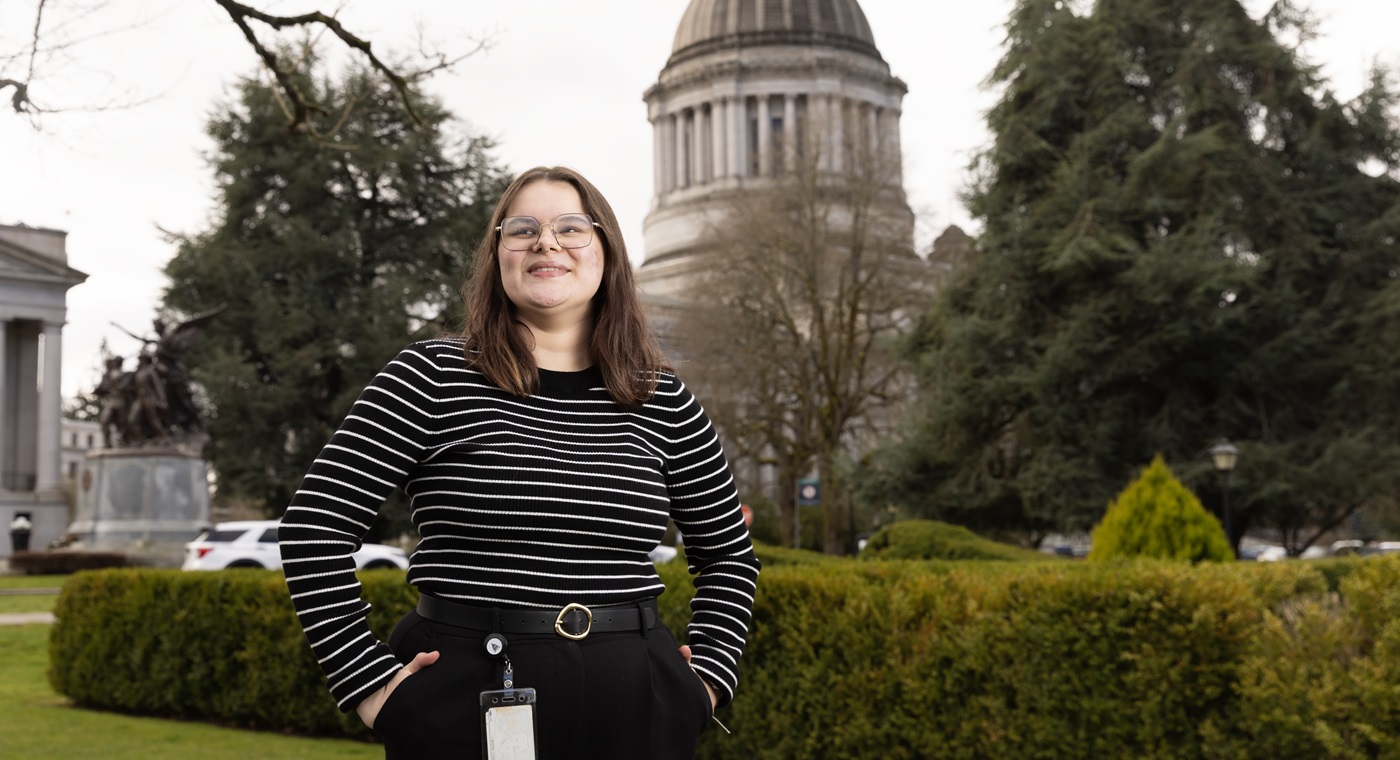 Cady Wood, dressed in a black and white striped shirt, and black pants with a badge attached to her belt, stands with her hands in pockets outside, looking just beyond the camera. Behind her are bushes, trees and the large white and grey Washington State capitol building.