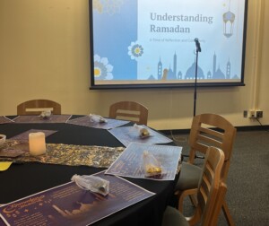 Blog - Iftar 03 A picture of a round, set table with a black tablecloth, purple placemats that also describe Ramadan and a candle in the middle. Behind the table is a screen on the wall, with a slide that reads 'Understanding Ramadan'
