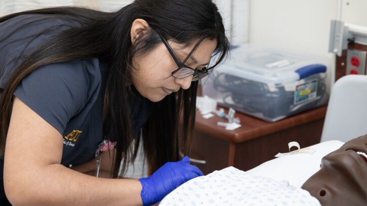 A PLU nursing student in dark grey scrubs leans over a simulated patient in a hospital bed in PLU's Nursing Simulation Clinical Center