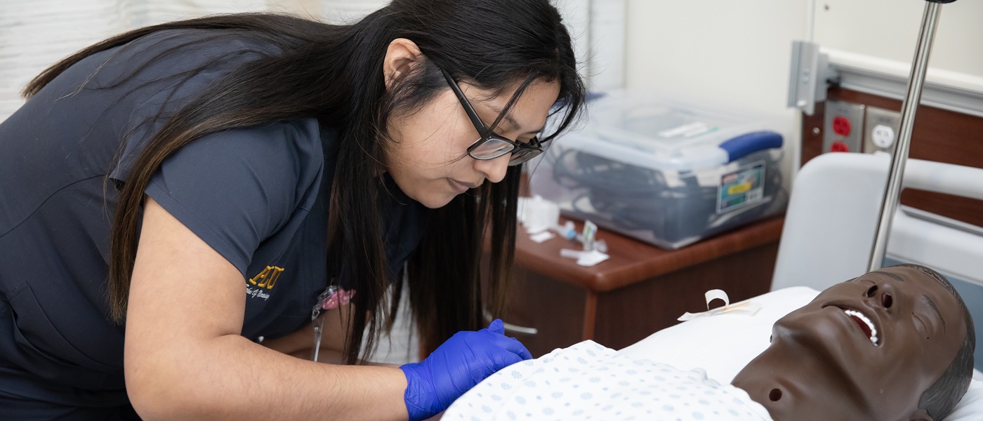 A PLU nursing student in dark grey scrubs leans over a simulated patient in a hospital bed in PLU's Nursing Simulation Clinical Center