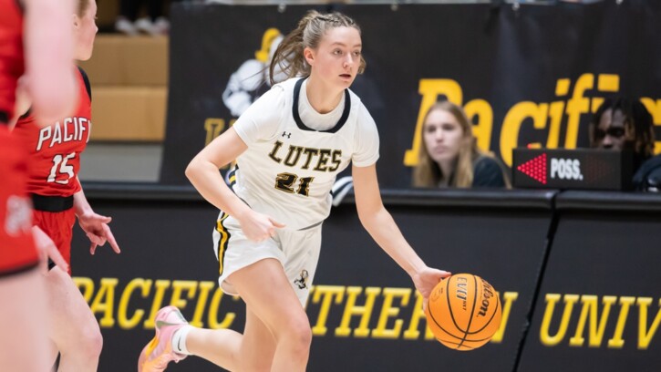 A PLU women's basketball player runs across the court with the ball. To the left are opposing players, and in the background to the right is the media box with currents students.