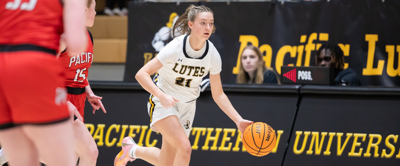 A PLU women's basketball player runs across the court with the ball. To the left are opposing players, and in the background to the right is the media box with currents students.