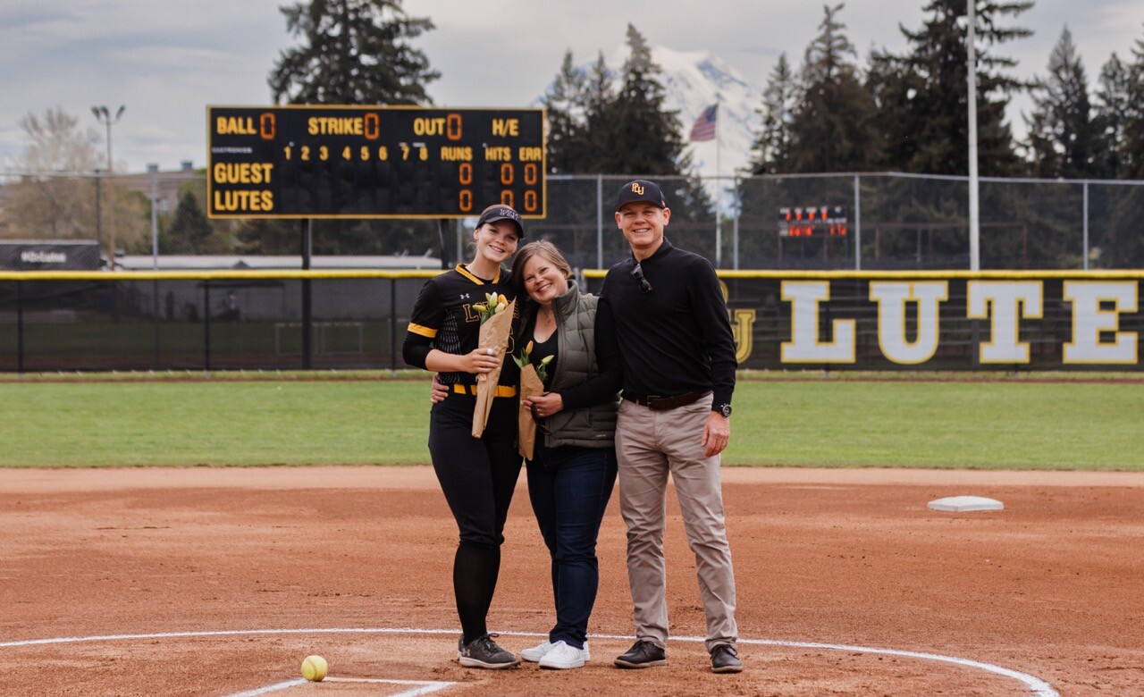 Molly House in her PLU softball uniform, holds a bouquet of flowers and poses with her parents on the PLU softball field.