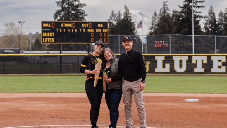 Molly House in her PLU softball uniform, holds a bouquet of flowers and poses with her parents on the PLU softball field.