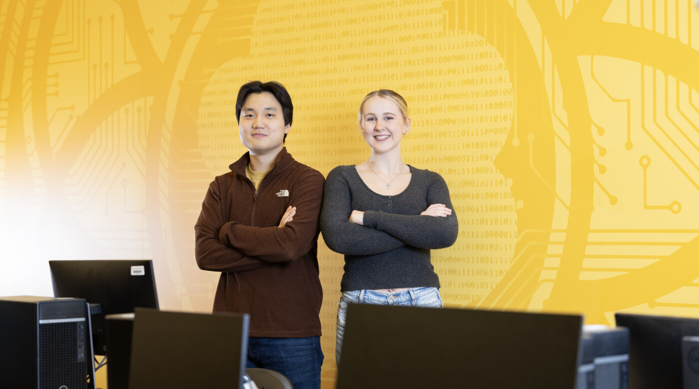 Andrew Lee and Stacie Spahr stand together with arms crossed in one of PLU's computer science labs. The room is brightly lit, and the wall behind them is painted PLU yellow with designs and computer science coding.