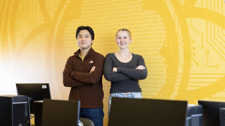 Andrew Lee and Stacie Spahr stand together with arms crossed in one of PLU's computer science labs. The room is brightly lit, and the wall behind them is painted PLU yellow with designs and computer science coding.