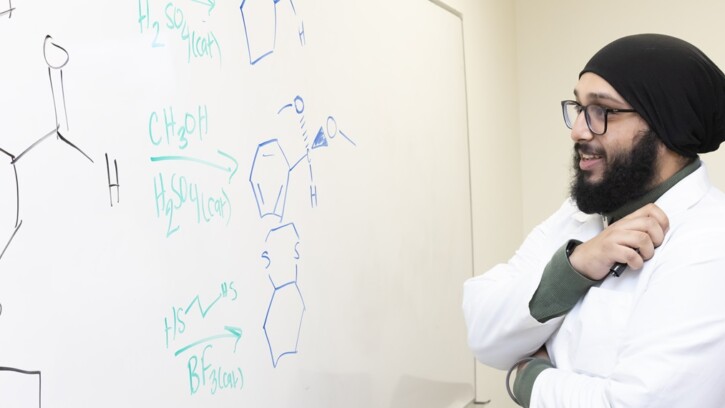 Chemistry and Religion double major Justin Singh hopes to become a doctor one day. Wearing a white lab coat, Justinpal stands at a white board with chemical molecules written on it.
