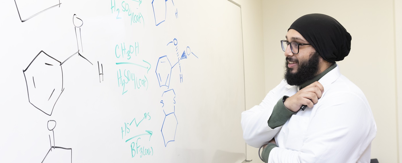 Chemistry and Religion double major Justin Singh hopes to become a doctor one day. Wearing a white lab coat, Justinpal stands at a white board with chemical molecules written on it.