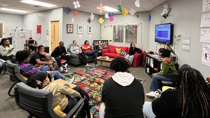 About a dozen students in PLU's Black Student Union sit in a circle in chairs and couches in PLU's Diversity Center during a meeting. Posters and framed artwork are on the walls, and colorful paper snowflakes hang from the ceiling.