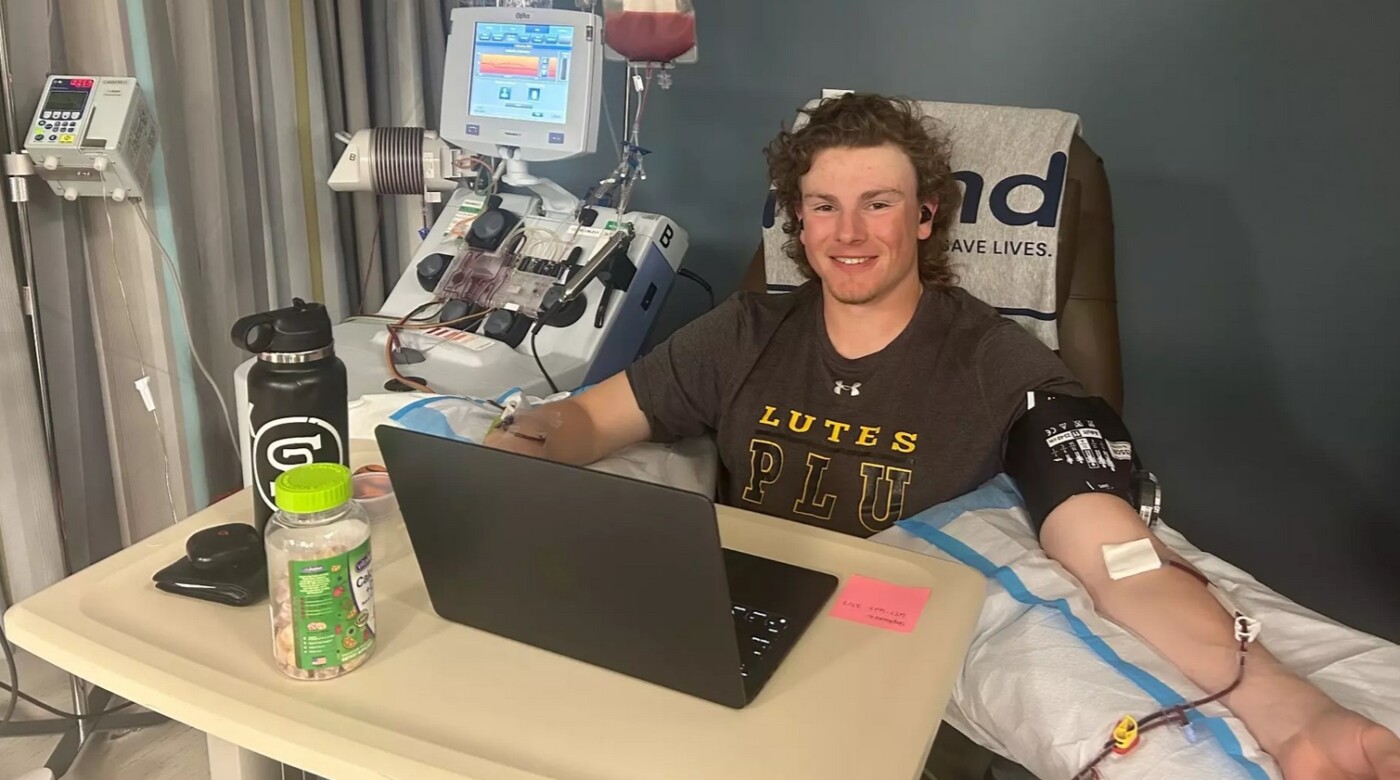 Carson Burke, in a PLU t-shirt, sits up in a hospital bed with his laptop open in front of him. His left arm is propped up on a pillow with an IV inserted, as he donates stem cells.
