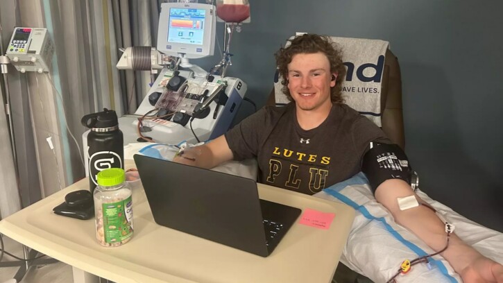 Carson Burke, in a PLU t-shirt, sits up in a hospital bed with his laptop open in front of him. His left arm is propped up on a pillow with an IV inserted, as he donates stem cells.