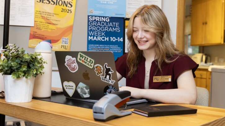 Gracie Hansen, in a maroon shirt with a gold name tag, sits at a desk with various things on it, including her open laptop with stickers. On the wall beside her are posters with information about campus events.