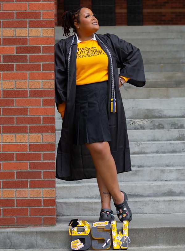Blog - Zoneice Grose 01 Social Work major Zoneice Grose stands on the grey staircase of PLU's Mary Baker Russell Music Center. She is leaning against a red brick pillar and looking off to her left. She's wearing open graduation robes and cords, with a yellow shirt that reads 'Pacific Lutheran University' in all caps and a short black skirt. At her feet are the letters 'BSW' decorated in black and yellow.