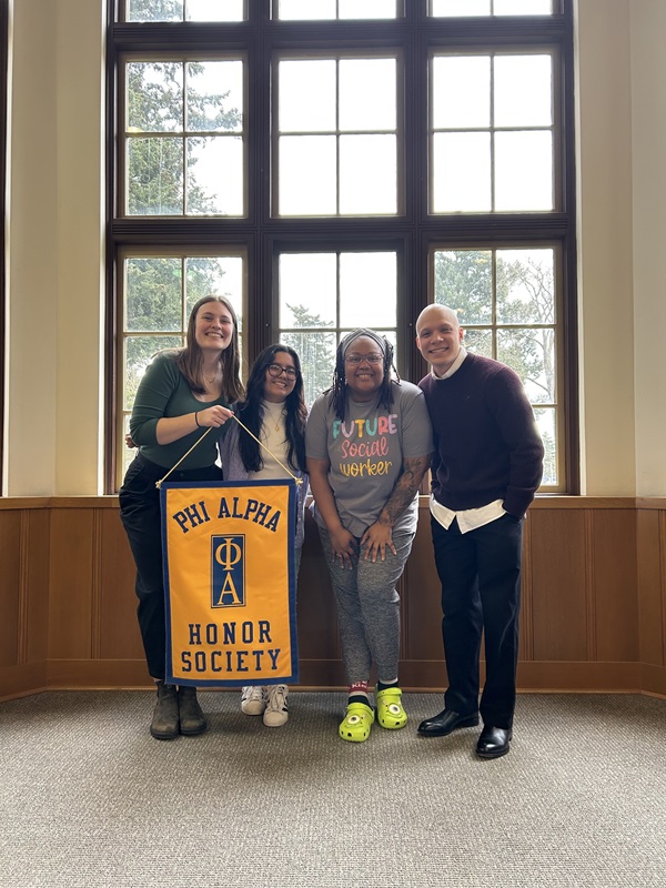Blog - Zoneice Grose 02 Zoneice and three other students pose together in front of a large sunlit window in one of PLU's academic buildings. One of the other students is holding a large banner that reads 'Phi Alpha Honor Society'.